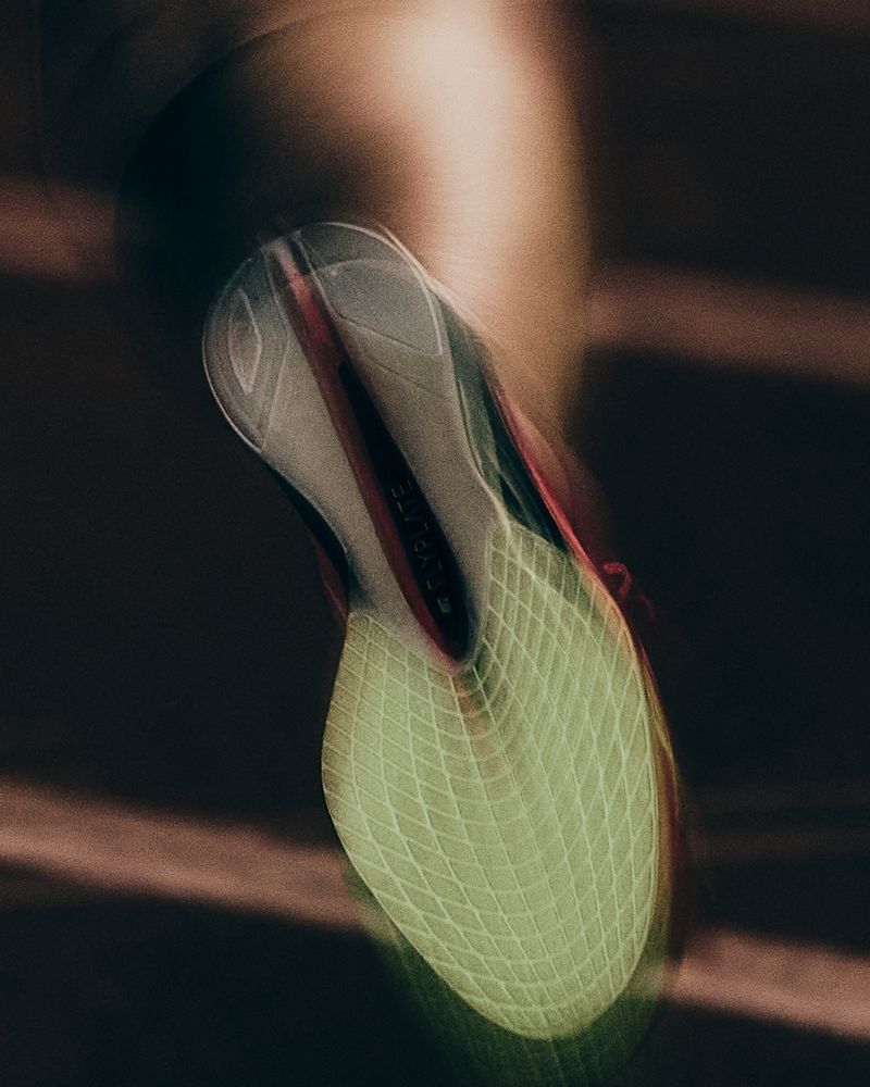 Close-up of a running shoe sole in motion with visible motion blur captured from below against a dark track.