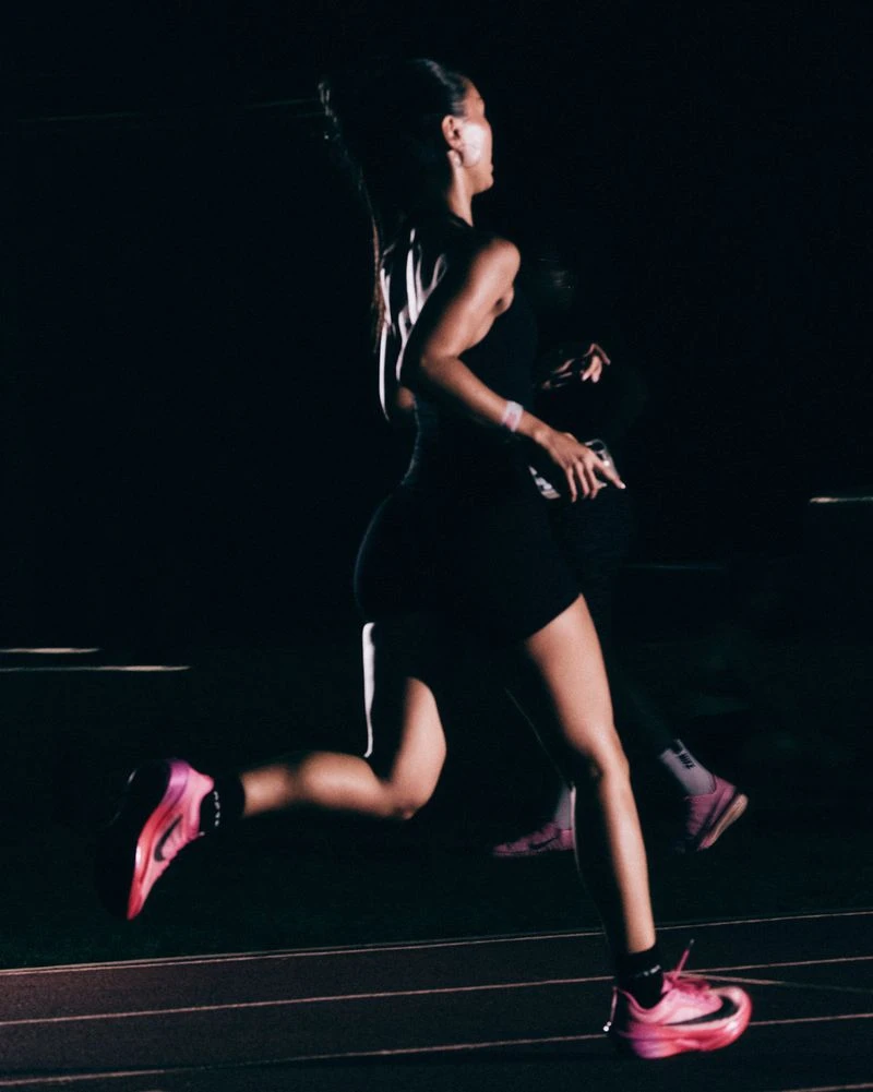 Female runner wearing dark athletic gear and pink running shoes sprinting on a track at night for Nike After Dark.