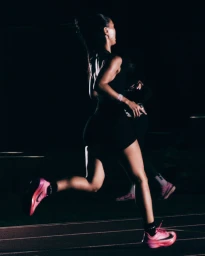 Female runner wearing dark athletic gear and pink running shoes sprinting on a track at night for Nike After Dark.