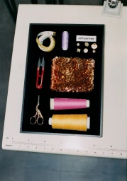Overhead flat lay of sewing supplies including thread spools, scissors, measuring tape, and fabric swatch on black tray