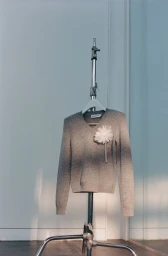 Grey v-neck sweater with a white flower embellishment hanging on a metal stand against a pale wall.