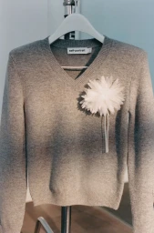 Grey knitted v-neck sweater on a white hanger with a white fabric flower pin attached to the chest