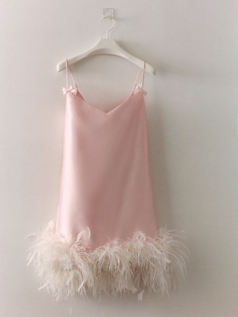 A pink slip dress with feather trim hanging on a white hanger against a plain white wall.