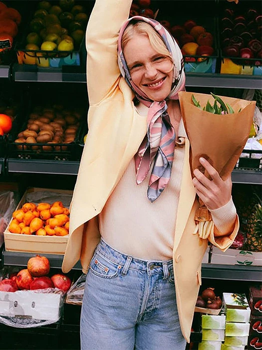 Smiling model wearing a silk headscarf and yellow blazer holding a bouquet of flowers in a grocery store market aisle