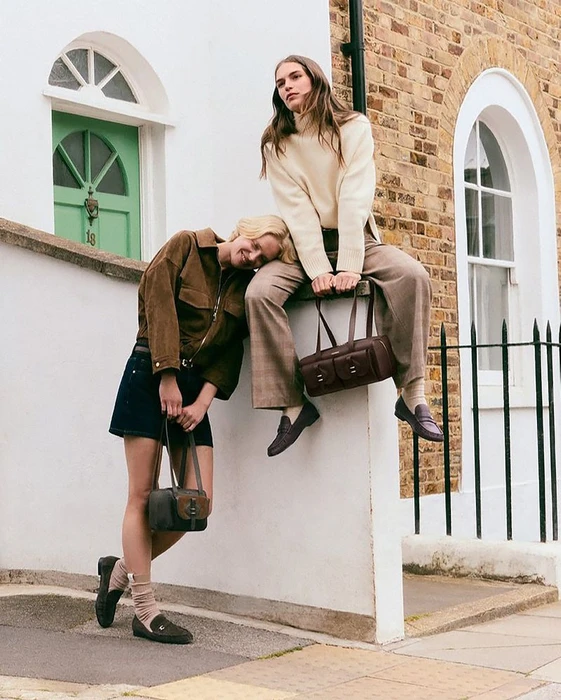 Two female models posing outdoors against a white building with brick trim, holding leather handbags.