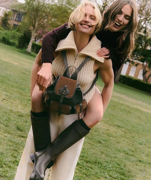 Two smiling models outdoors on a grassy lawn, one giving the other a piggyback ride wearing a cream knit vest and leather backpack