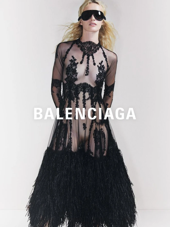 Model wearing a sheer black lace gown and dark sunglasses against a plain white studio background.