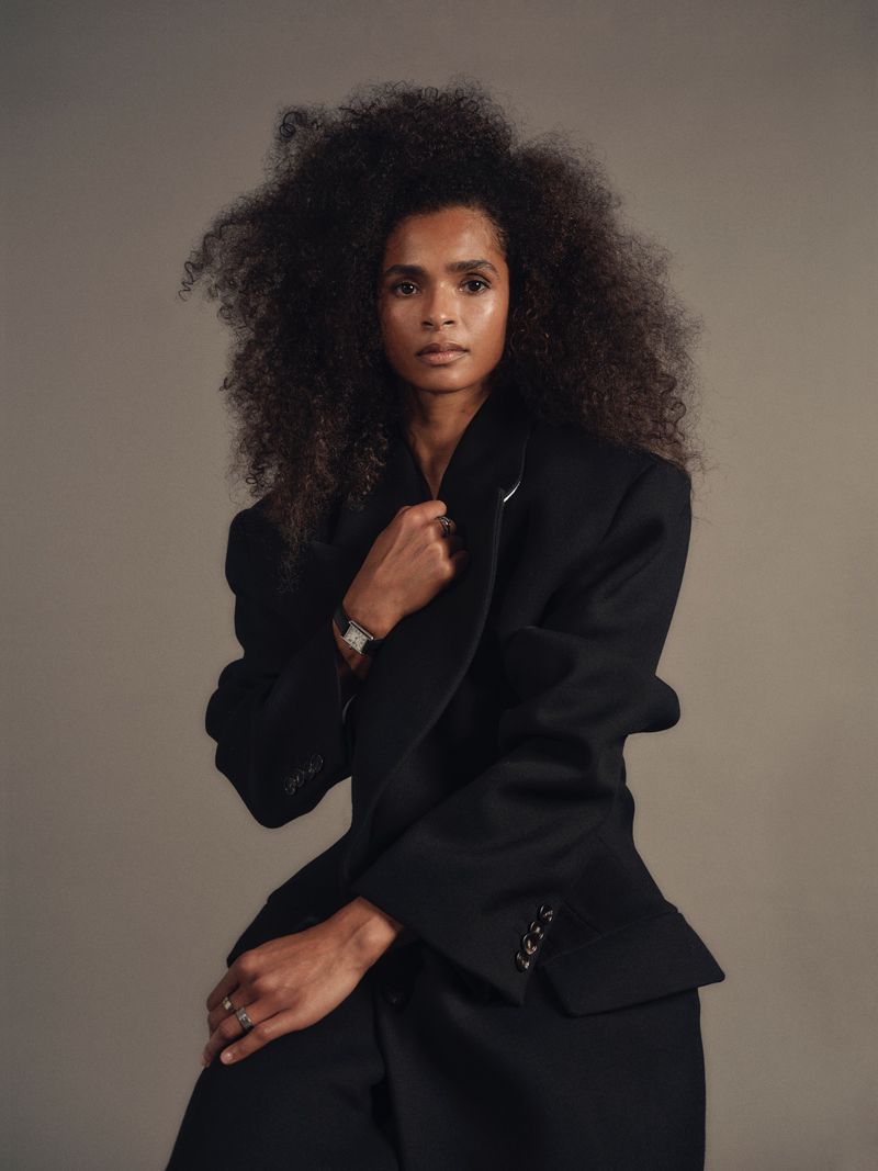 Portrait of Ramla Ali with voluminous dark curly hair wearing an oversized black blazer against a neutral grey studio background.