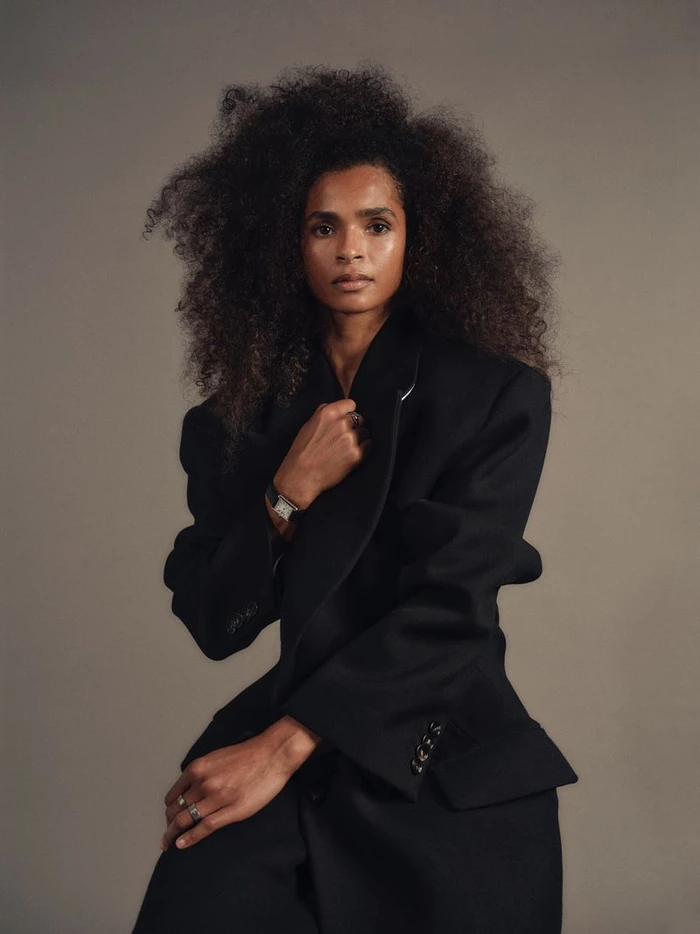 Portrait of Ramla Ali with voluminous dark curly hair wearing an oversized black blazer against a neutral grey studio background.