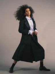 Ramla Ali posing in a dark blazer, striped shirt, and pleated skirt against a grey background for Elle UK.