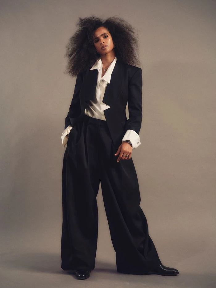 Ramla Ali standing in a black suit and white shirt against a neutral backdrop for an Elle UK editorial portrait.
