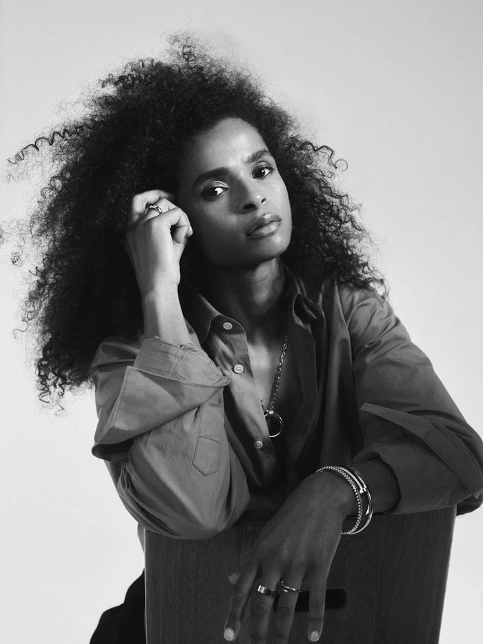 Black-and-white studio portrait of Ramla Ali wearing a collared button-down shirt, rings, and bracelets against a plain light background