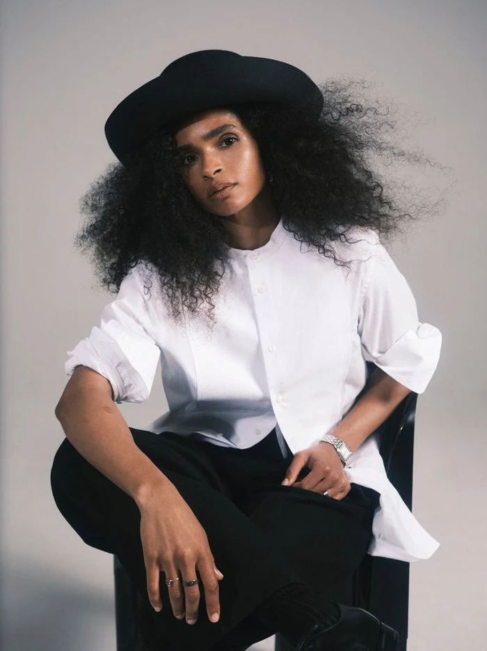 Athlete Ramla Ali seated wearing a black hat, white button-down shirt, and black trousers against a neutral background.