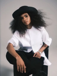 Athlete Ramla Ali seated wearing a black hat, white button-down shirt, and black trousers against a neutral background.