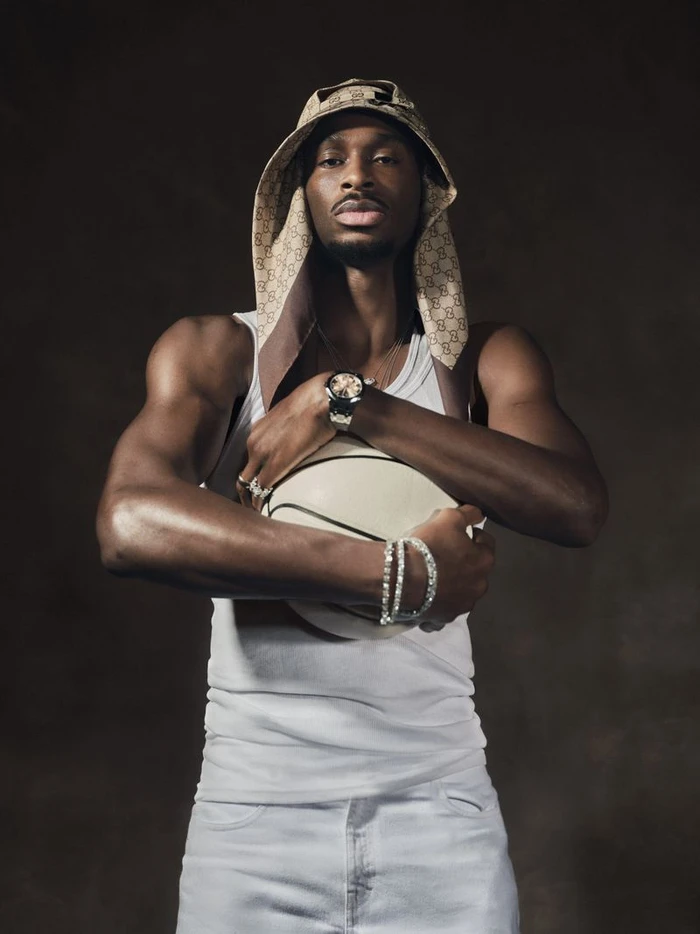Shai Gilgeous-Alexander holding a basketball, wearing a white tank top and patterned bucket hat against a dark backdrop