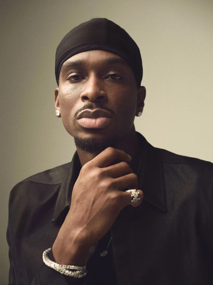 Shai Gilgeous-Alexander wearing a black shirt and skullcap, resting his hand under his chin against a neutral studio backdrop.