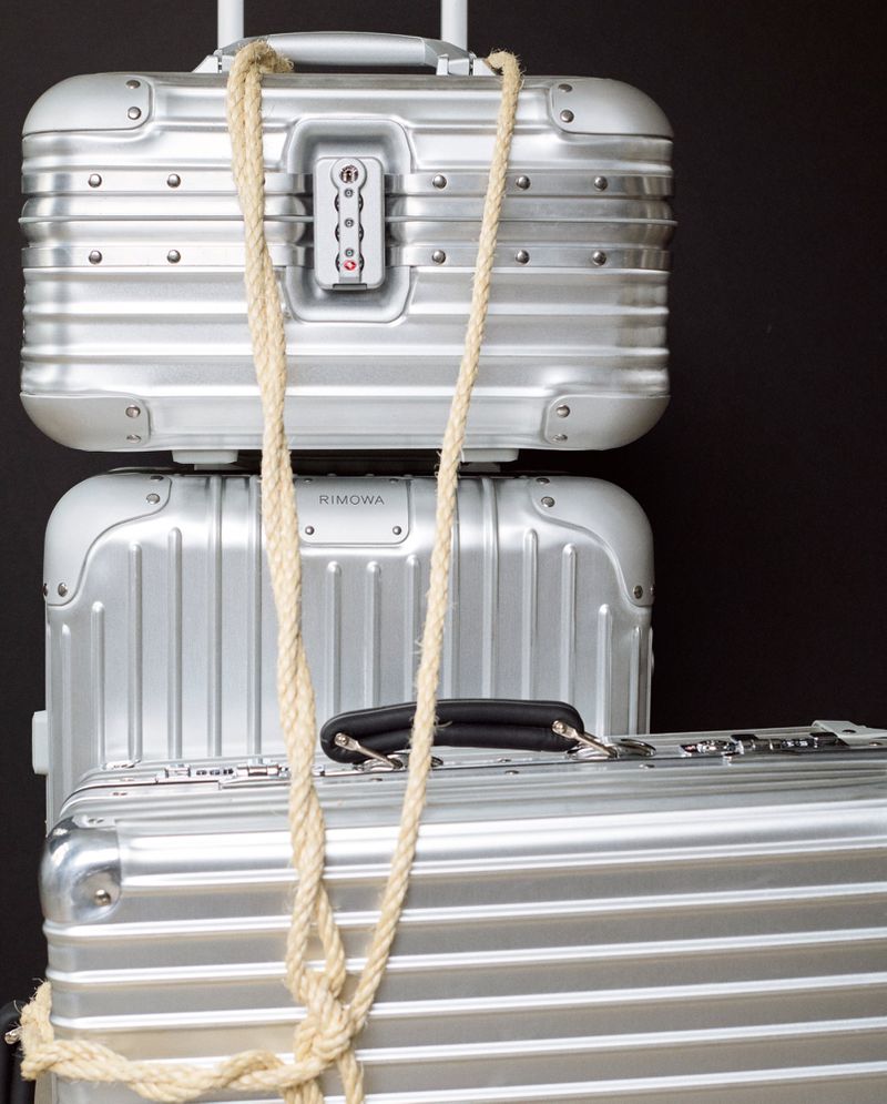 Stack of three Rimowa aluminum suitcases secured with a tan rope against a dark backdrop, evoking travel and premium luggage.