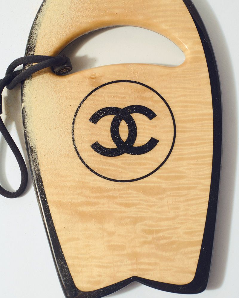Close-up of light brown surfboard with a black 'CC' logo design and a black trim and black leash on a white background.
