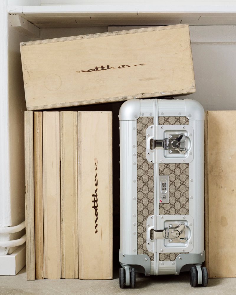 Silver Gucci suitcase on wheels next to wooden boxes. The suitcase is patterned with interlocking G logos on the side.