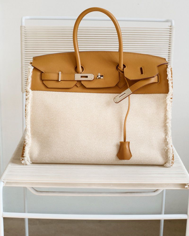 Tan leather handbag with white canvas and fringe detail sits on a white chair against a plain background in a product shot.