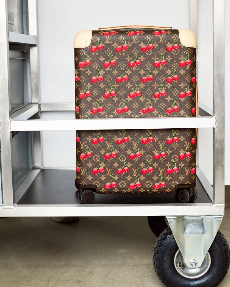 Brown Louis Vuitton suitcase with cherry pattern on a metal luggage cart in a bright setting, showcasing luxury travel accessories.