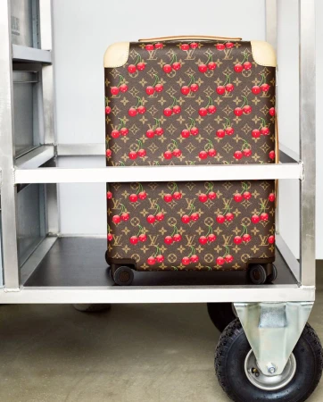 Brown Louis Vuitton suitcase with cherry pattern on a metal luggage cart in a bright setting, showcasing luxury travel accessories.