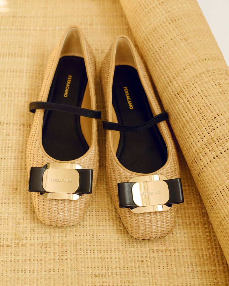 Woven beige ballet flats with a black strap and a gold buckle, displayed on a textured beige surface, elegant and fashionable.