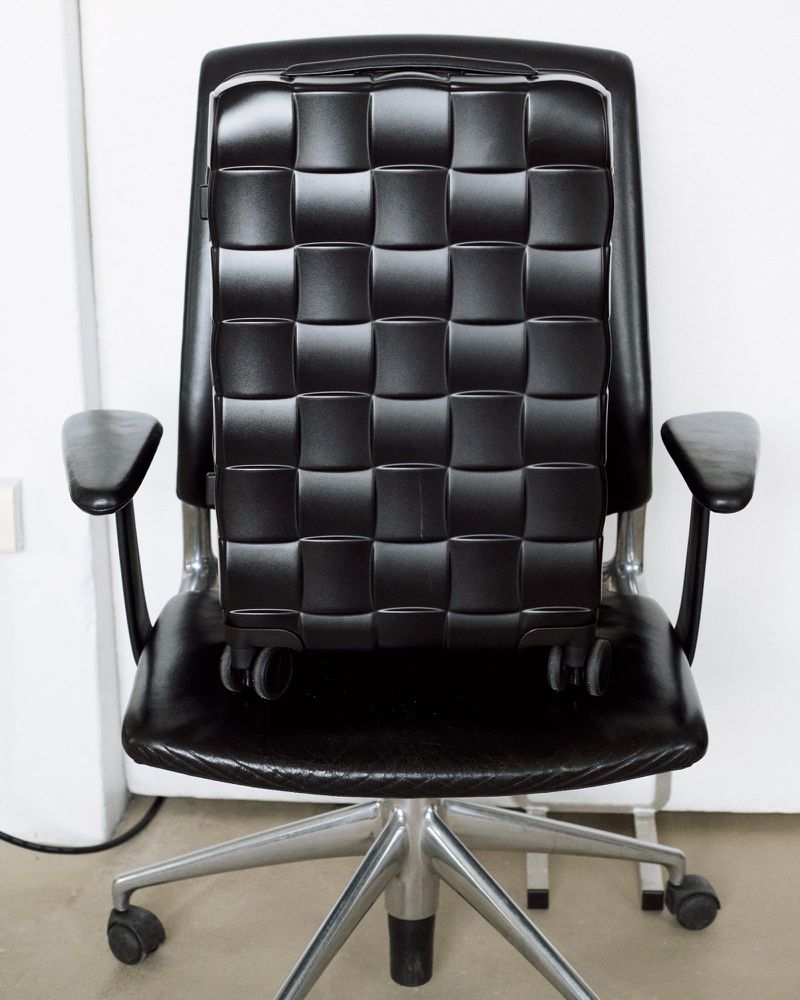 Black leather office chair with woven pattern back, set against a plain white wall in a well-lit interior.