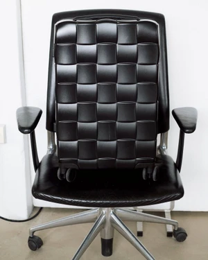 Black woven suitcase resting upright on a black leather office chair against a plain white background.