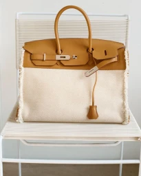 Canvas and tan leather top-handle handbag with frayed side trims resting on a minimalist white cord chair.