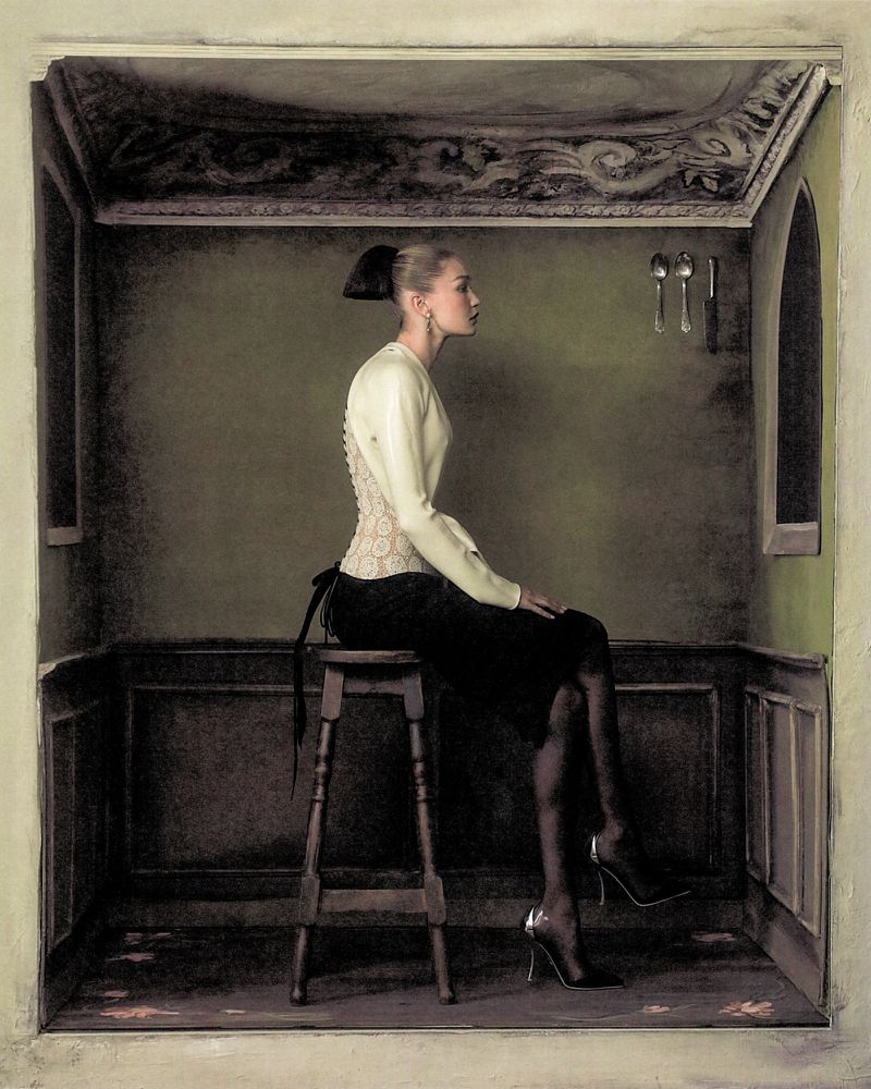 Gigi Hadid seated on a wooden stool inside a dark green room holding cutlery against the wall for WSJ Magazine Queen of Couture.