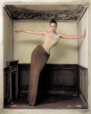 Model Gigi Hadid posing inside a painted architectural box set wearing a structured beige bodice and textured brown skirt.