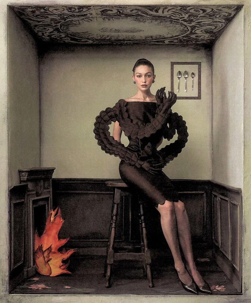 Gigi Hadid seated on a wooden stool inside a stylized room set wearing a structured black dress