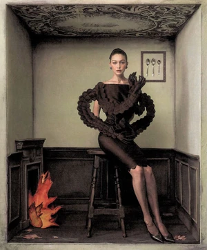 Gigi Hadid seated on a wooden stool inside a stylized room set wearing a structured black dress