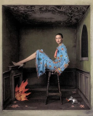 Gigi Hadid in a blue embroidered dress seated on a stool inside a dark room with a fireplace and painted ceiling