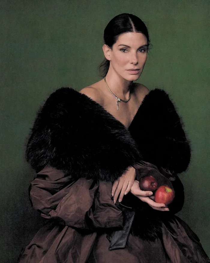 Actor Sandra Bullock holding two red apples while wearing a black fur stole and dark dress against a textured green background