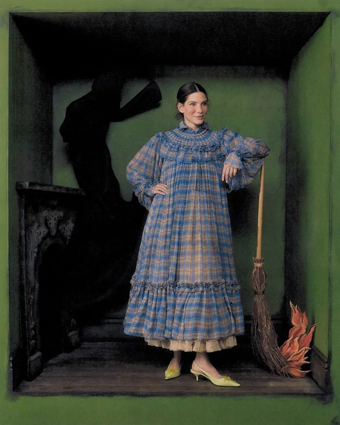 Sandra Bullock in a blue plaid smocked dress posing inside a dark green set with a wooden broom and a decorative fireplace