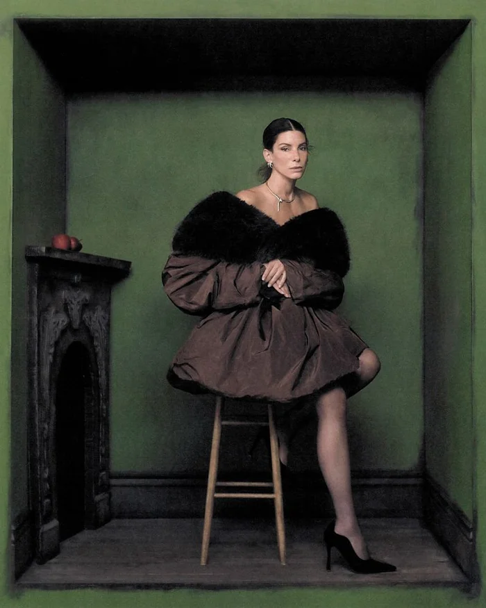 Sandra Bullock seated on a stool inside a green room wearing a brown puff coat and black high heels for Interview Magazine