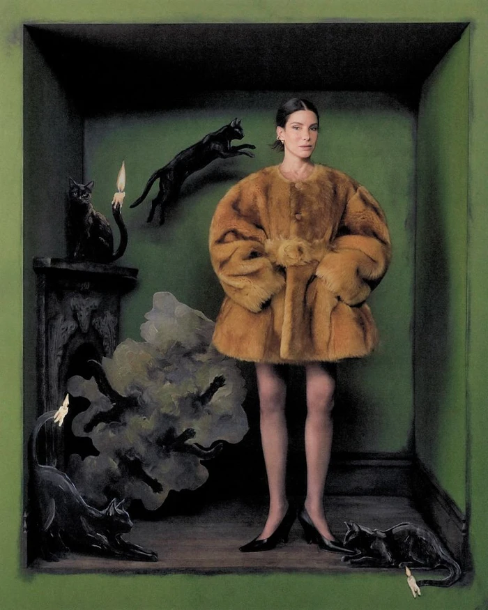 Sandra Bullock wearing an oversized caramel fur coat inside a dark green niche surrounded by black cats and candles.