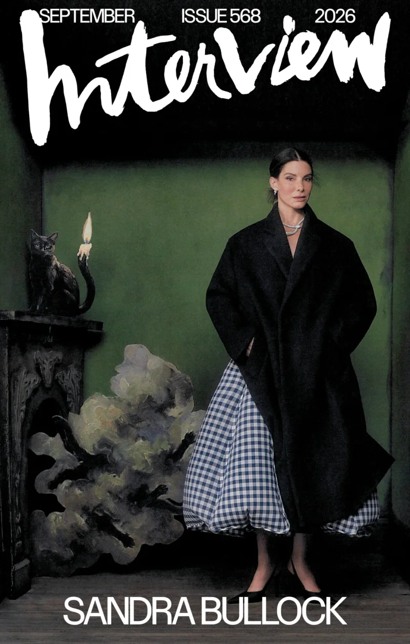 Sandra Bullock wearing a dark coat and checkered skirt standing in a green room with a fireplace.