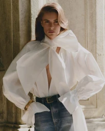 Model wearing a white blouse with a large bow and blue jeans standing against a stone architectural pillar.