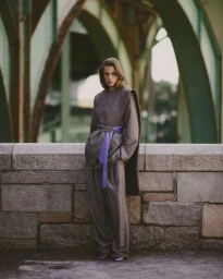Model Lydia-Angel in a grey wool suit with a purple belt standing before a stone wall and arched bridge structure.