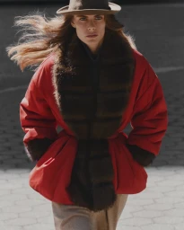 Model wearing a red coat with a dark fur collar and a hat outdoors against an urban street background.