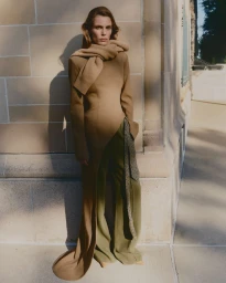 Model standing outdoors against a stone building exterior wearing a camel dress and scarf with olive green trousers.