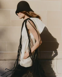 Model wearing a black cloche hat, cream hooded top with black braided trim, and dark pants against a stone wall.