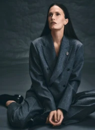 Model seated wearing a grey flannel double breasted suit against a dark textured backdrop for Harper's Bazaar Germany.
