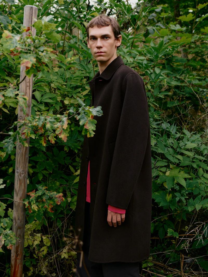 Model Aleksandr Gordeev wearing a dark brown coat in a lush green woodland setting for Arket.