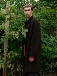 Model Aleksandr Gordeev wearing a dark brown coat in a lush green woodland setting for Arket.