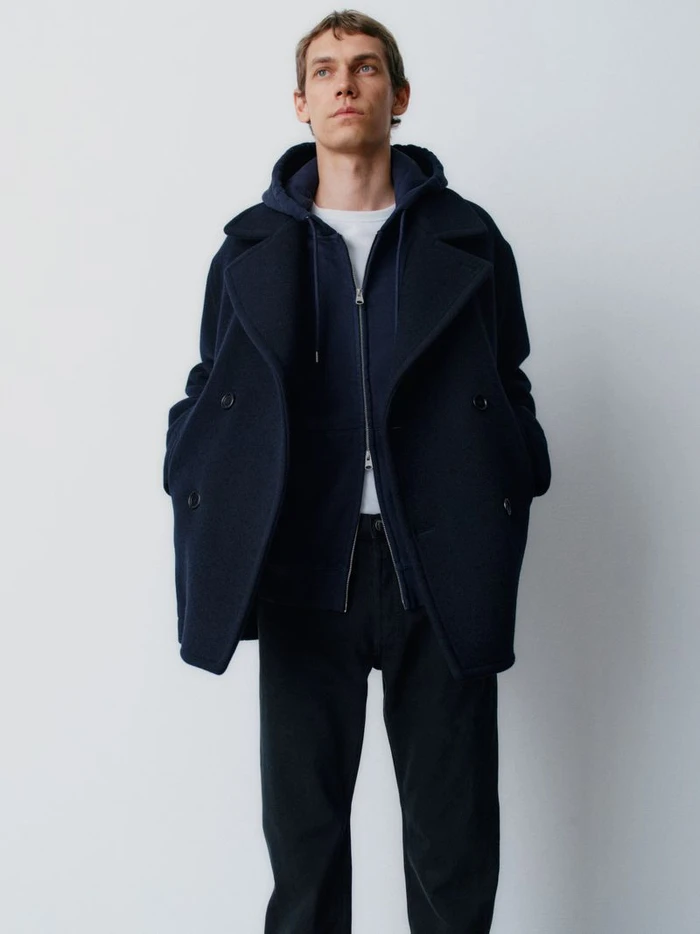 Male model wearing a navy blue peacoat over a zipped hoodie against a plain light grey backdrop