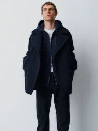 Male model wearing a navy blue peacoat over a zipped hoodie against a plain light grey backdrop
